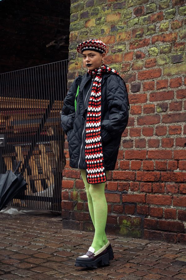 Mulher morena com cabelo raspado posando para foto nas ruas da Dinamarca. Ela usa um casaco estilo puffer, meia calça verde, mocassim preto e branco e um conjunto de cachecol + boina com uma padronagem preta, branca e vermelha.