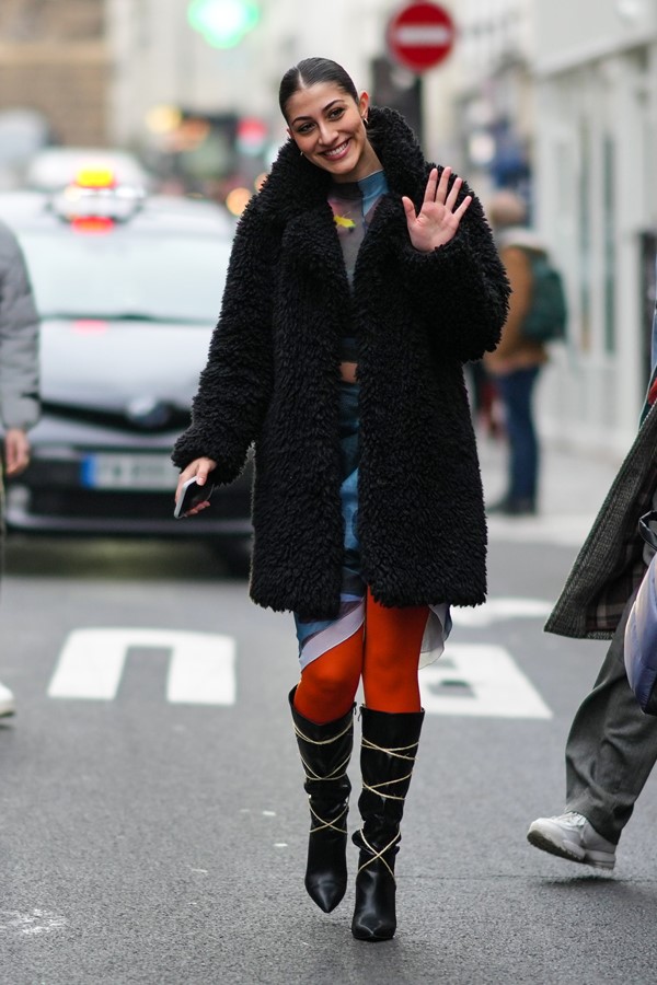 Mulher branca com cabelo preto amarrado andando pelas ruas de Nova York, nos Estados Unidos. Ela usa brincos de ouro, uma camiseta cinza claro/azul/marrom/amarelo de gola alta, uma saia envelope cinza claro/azul/marrom/amarelo, um casaco longo preto macio, meia-calça laranja, saltos pontiagudos de couro preto brilhante botas de joelho com cadarços bege.