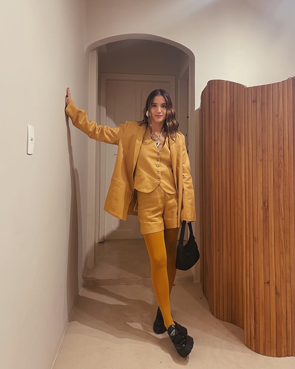 Mulher branca, com cabelo ondulado castanho claro, posando para foto no corredor de um apartamento. Ela usa um conjunto amarelo de shorts, colete e blazer, e veste, ainda, uma meia calça também amarela. Segura uma bolsa preta e usa um sapato mocassim também preto.