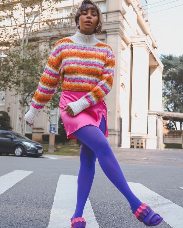 Mulher negra, com cabelo curto e liso, posando para foto nas ruas de São Paulo, mais precisamente na faixa de pedestre. Ela usa um casaco de tricô lsitrado nas cores branco, laranja e bosa, uma sala rosa, uma meia-calça roxa e uma sandália aberta de salto também rosa e lilás.