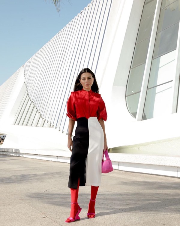 Mulher branca, com cabelos castanhos e lisos, posando para foto em museu do Rio de Janeiro. Ela usa uma blusa e meia calça vermelha e uma saia preta e branca, além de uma bolsa rosa.