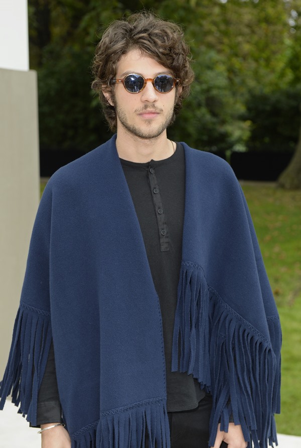 O ator Chay Suede, um homem branco com cabelo médio castanho, posando para foto na entrada do desfile da Burberry, na Inglaterra. Ele usa um óculos escuro, camista e calça pretos e um poncho azul por cima.