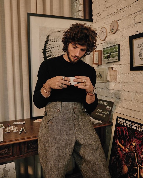 O ator Chay Suede, um homem branco com cabelo médio castanho, tomanco café na sala de casa, encostado em uma mesa de madeira. Ele veste uma blusa de manga longa preta e uma calça cinza.