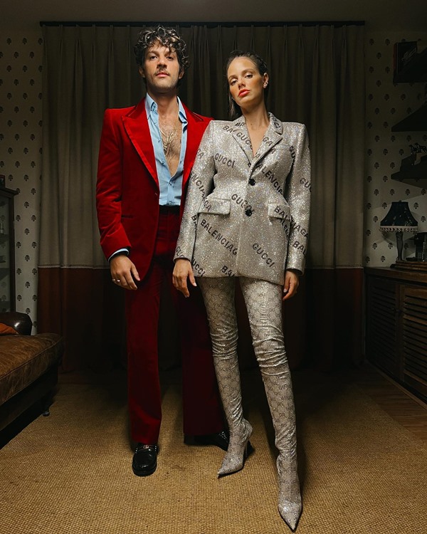 O ator Chay, um homem branco com cabelo e barba na cor castanha, posando para foto com sua esposa, a modelo Laura Neiva, uma mulher branca com cabelo liso castanho. Ele usa um terno vermelho de veludo com uma camisa azul aberta e ela usa um blazer prateado com uma da mesma cor.