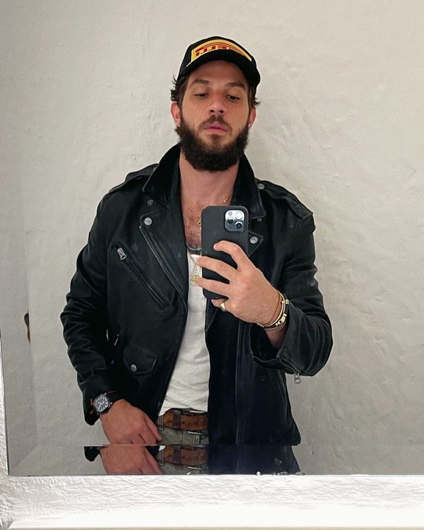 O ator Chay, um homem branco com cabelo e barba na cor castanha, posando para foto na frente do espelho com o celular. Ele usa uma regata branca, calça jeans, uma jaqueta de couro e um boné preto.