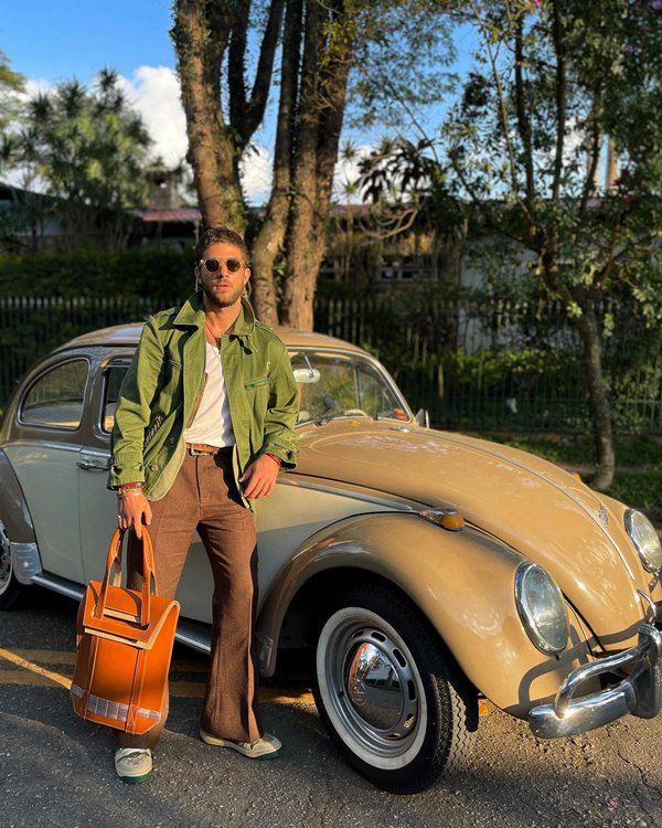 O ator Chay, um homem branco com cabelo e barba na cor castanha, posando para foto na frente de um fusquinha bege. Ele usa uma blusa branca, calça marrom, uma jaqueta verde militar e uma bolsa caramelo.