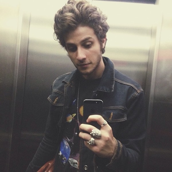 Chay Suede, um homem branco com cabelo castanho, posando para foto na frente do espelho em um elevador. Ele usa uma camiseta preta, com estampa do sistema solar, e uma jaqueta jeans.