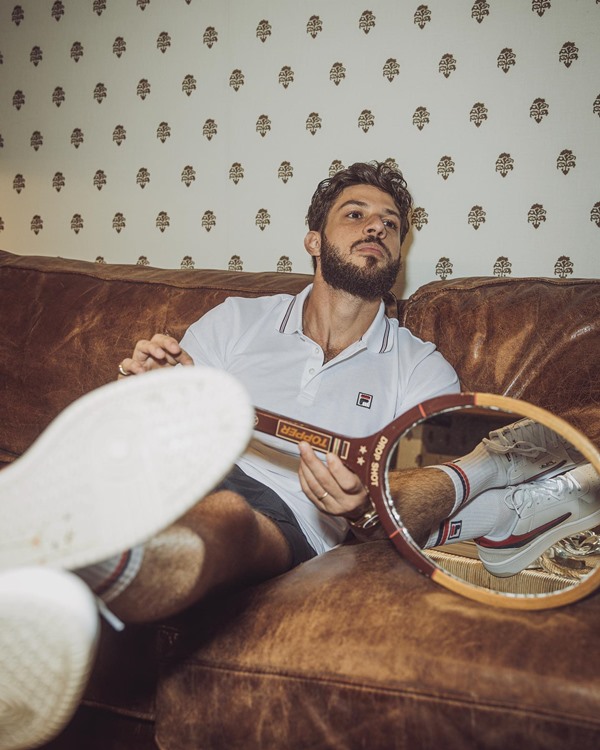 O ator Chay, um homem branco com cabelo e barba na cor castanha, posando para foto em um sofá de couro marrom. Ele usa uma camisa polo branca, short azul escuro, meias e tênis branco e segura uma raquete de tênis nas mãos.