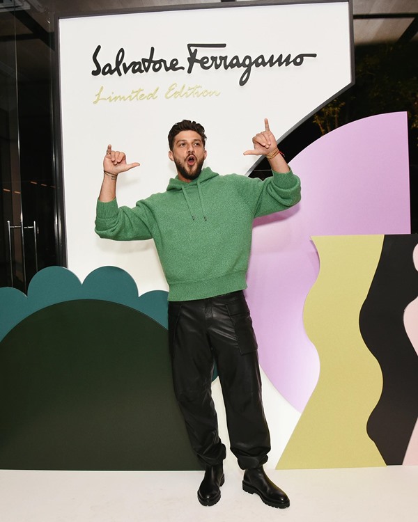 O ator Chay, um homem branco com cabelo e barba na cor castanha, posando para foto em um evento da marca Salvatore Ferragamo. Ele usa uma calça preta de couro, um moletom verde e uma bota preta.