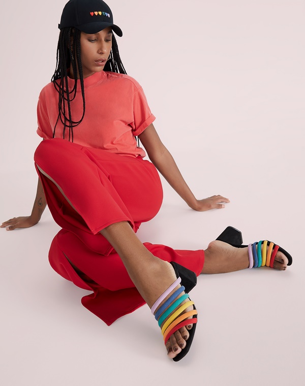 Mulher negra com cabelo trançado longo posando para foto em um fundo branco. Ela usa uma camiseta e uma calça vermelha, boné preto e uma sandália de salto com tiras nas cores do arco-íris. As peças são da Amaro.