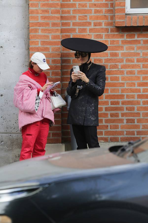 Mulher usa look vermelho com boné branco ao lado de homem com visual preto com alfaiataria e chapéu