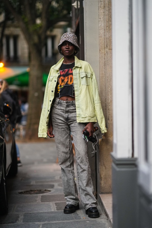 Mulher negra com cabelo curto posando para foto na rua. Ela veste um chapéu estilo pescador com estampa de onça, brincos e um colar de corrente dourados, uma camiseta preta com a logo da banda AC/DC, uma jaqueta jeans amarelo pálido, calça jeans preta flare desbotada, bota coturno preta da Dr Martens.