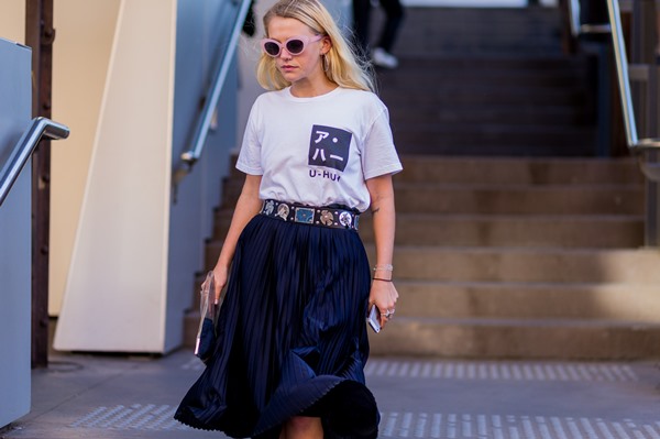 Mulher branca e com cabelo liso louro, andando pela rua, com óculos escuros de armação lilás. Ela usa uma camiseta branca, com um desenho de símbolos japoneses pretos, uma saia plissada preta e um cinto de taxas.