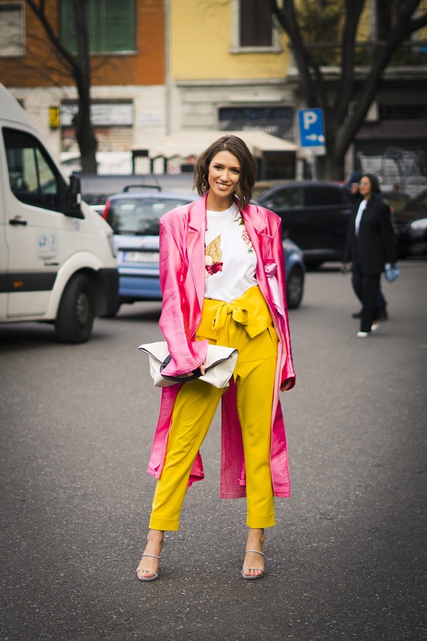 Mulher branca com cabelo castanho curto, ondulado, posando para foto nas ruas durante a Semana de Moda de Milão, na Itália. Ela veste uma camiseta branca, calça cenoura na cor amarela, um sobretudo rosa cintilante e um salto alto fino brilhante.