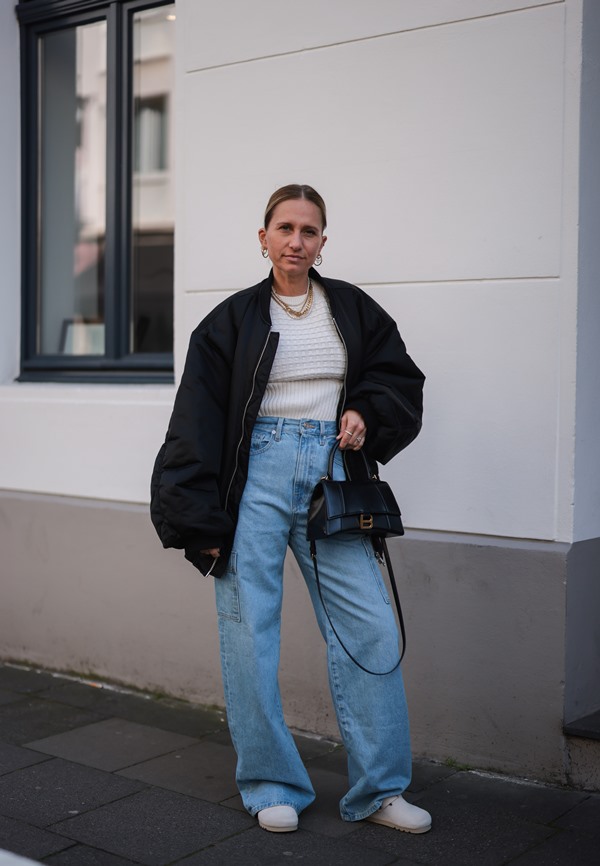 Mulher branca com cabelos louros amarrados posando para foto na rua. Ela usa uma regata branca, colar de corrente dourado, casaco preto, calça jeans e bolsa preta da Balenciaga.