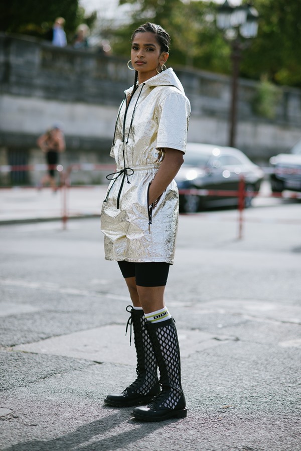 Mulher negra com cabelo trançado posando para foto na rua durante a Semana de Moda de Paris. Ela usa um macacão curto, dourado com detalhes pretos, meia branca longa da Dior e um coturno preto longo.