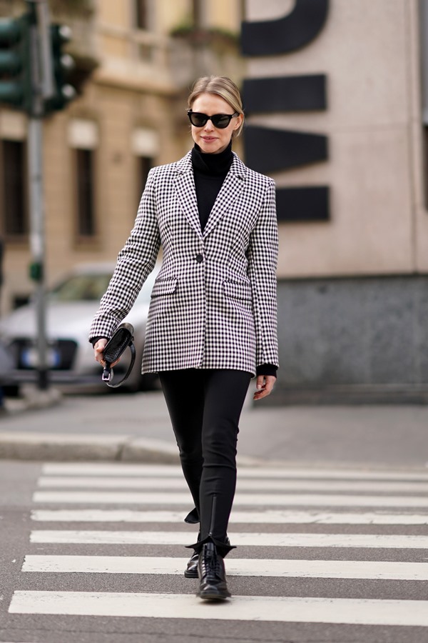 Mulher branca com cabelo louro e liso, preso em rabo de cavalo baixo, andando pelas ruas de Milão durante a Semana da Moda. Ela veste uma blusa de manga longa e gola alta preta, com calça preta, coturno de couro e um blazer quadriculado, estilo vichy, também preto e branco. Usa também um óculos escuros e segura, na mão direita, uma bolsa pequena.