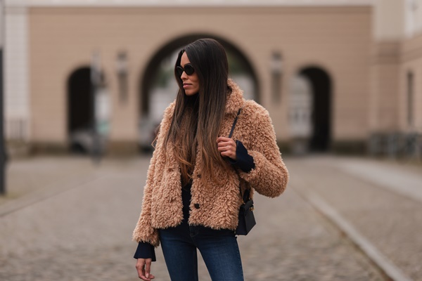 Mulher morena, com cabelos lisos e cumpridos, posando para foto nas ruas de Paris. Ela usa óculos escuros, blusa preta de manga longa, casaco teddy peludo marrom e calça jeans. Leva, no braço esquerdo, uma bolsa de corrente também preta.