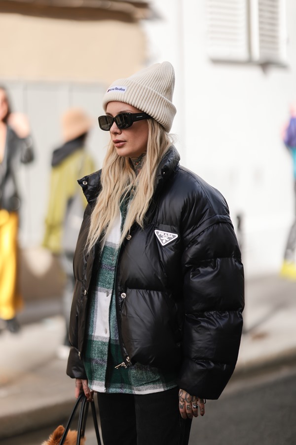 Mulher branca com cabelos cumpridos lisos, e louros, posando para foto na rua durante a Semana de Moda de Paris. Ela usa um óculos escuros e uma touca bege; um casaco de frio listrado branco e verde e, por cima, um casaco estilo puffer com a logo da Prada.