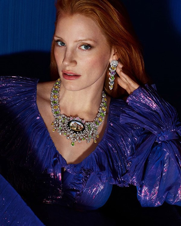 A atriz Jessica Chastain, uma mulher branca com cabelosos ruivos lisos e longos, posando para campanha publicitária de joias da marca Gucci. Na foto, ela usa um vestido azul com mangas bufantes, e detalhes brilhosos, e um colar e um brinco de diamantes com outras pedras preciosas.