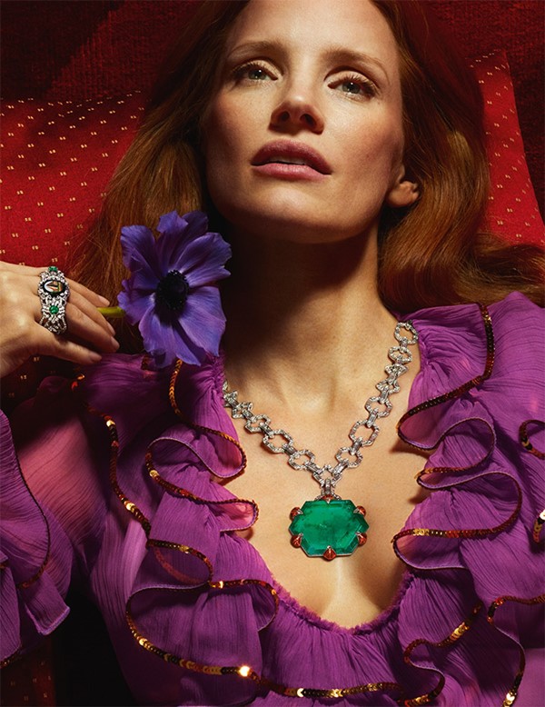 A atriz Jessica Chastain, uma mulher branca com cabelosos ruivos lisos e longos, posando para campanha publicitária de joias da marca Gucci. Na foto, ela usa um vestido roxo com babados na gola, e detalhes brilhantes, e um colar de corrente de ouro branco com uma esmeralda redonda no centro.