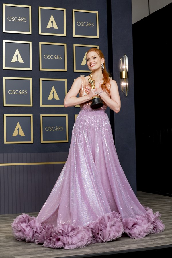 A atriz Jessica Chastain, uma mulher branca com cabelosos ruivos lisos e longos, na premiação Oscar de 2022, quando ganhou o troféu de Melhor Atriz. Na foto, ela usa um vestido da Gucci brilhante com plumas na ponta. Ele começa dourado e faz um degradê para o lilás.