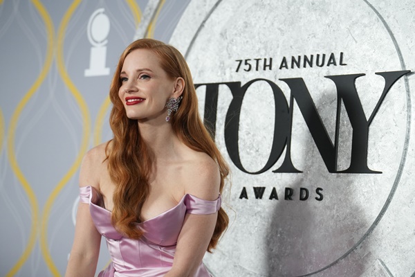 A atriz Jessica Chastain, uma mulher branca com cabelosos ruivos lisos e longos, na premiação anual Tony, voltada para quem faz musicais. Na foto, ela usa um vestido lilás de cetim com cauda da marca Gucci.