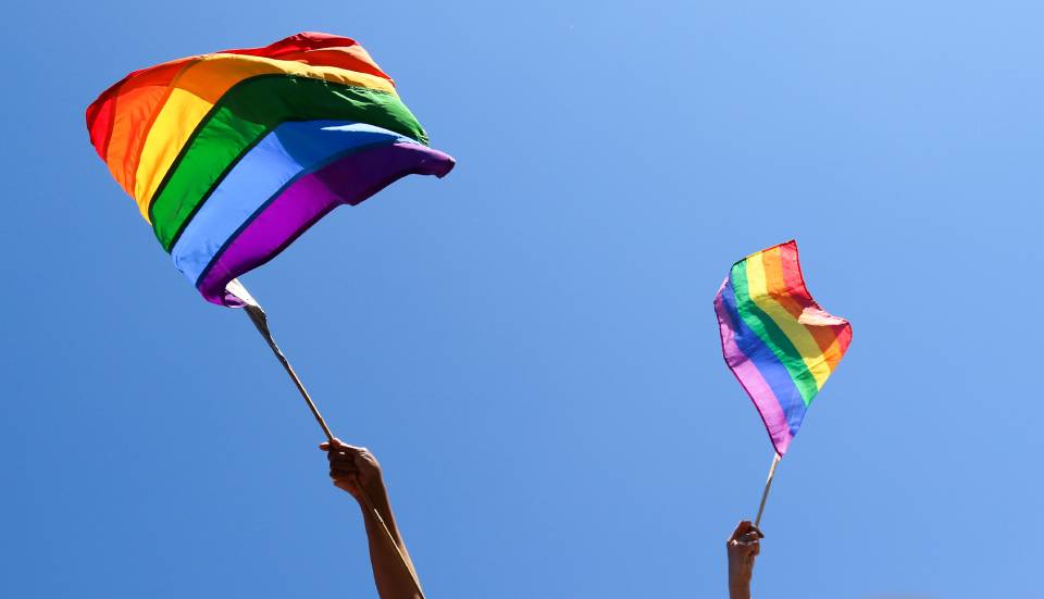 Pessoas balançam bandeiras LGBTQ+ de arco-íris no céu azul - Metrópoles