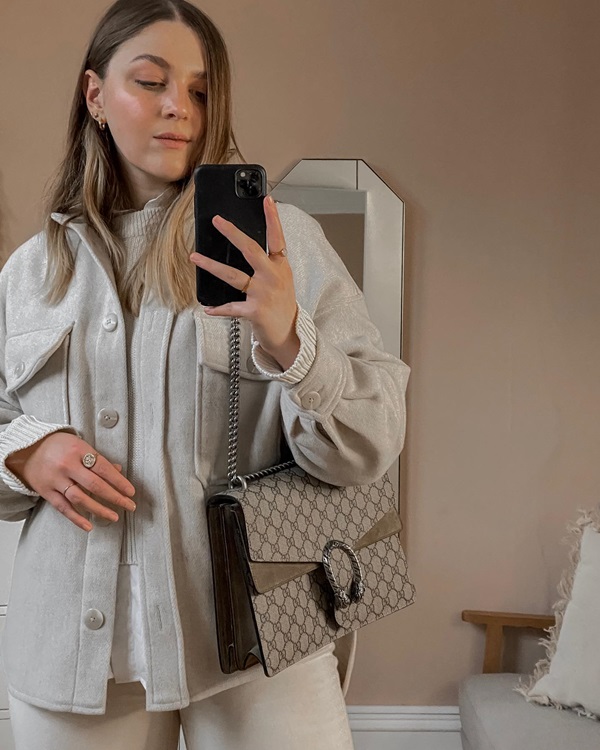 Mulher branca, com cabelo liso castanho claro, posando para foto em frente ao espelho e segurando o celular. Ela usa um suéter bege e uma jaqueta, também bege, por cima, e uma bolsa marrom da Gucci com correntes de prata.