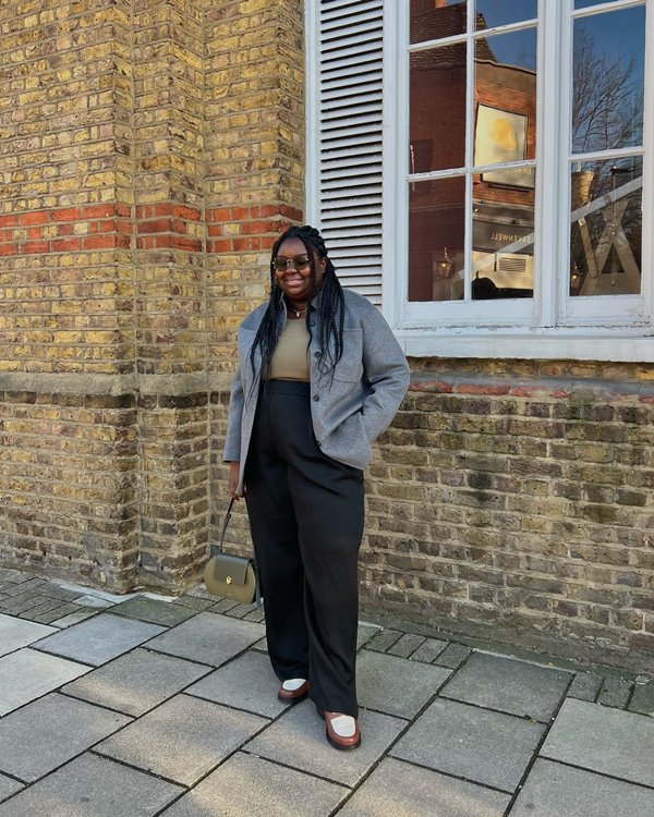 Mulher negra e jovem posando para foto nas ruas de Londres. Ela tem os cabelos trançados e cumpridos e veste uma blusa bege justa, uma jaqueta cinza e uma calça preta. Está de óculos escuros e usa ainda um sapato estilo oxford branco e marrom.