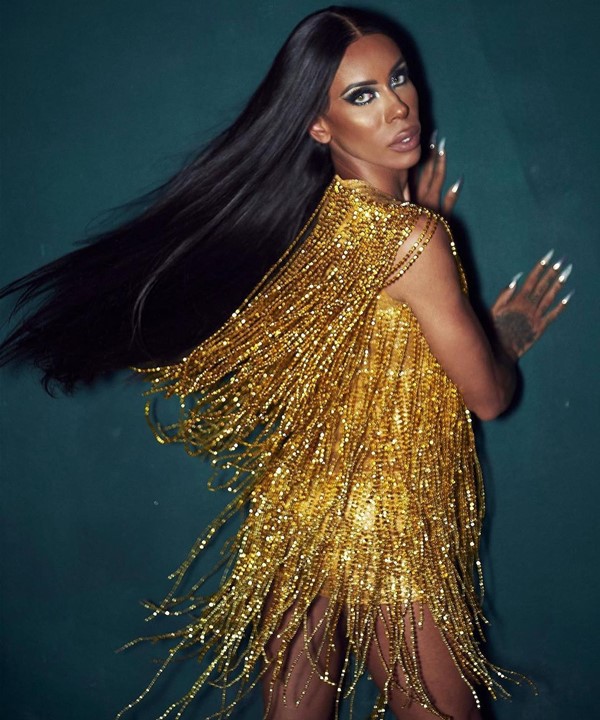 A drag queen Bianca Dellafancy, uma mulher de pele morena e cabelos pretos lisos e longos, posando para foto em um fundo cinza. Ela usa um vestido de franjas douradas do estilistas Walério Araújo.