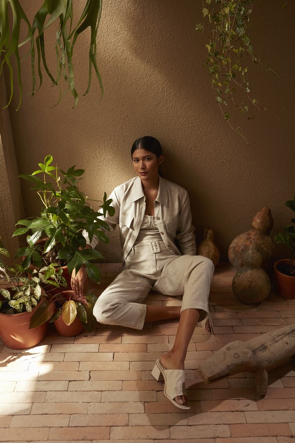 Foto de divulgação da nova coleção de roupas da marca Dafiti. Na foto, modelo posa com itens da etiqueta. É um mulher jovem, de pele morena e cabelos longos, lisos e pretos, que veste top, casaco e calça, tudo bege, em tecido de linho cru.