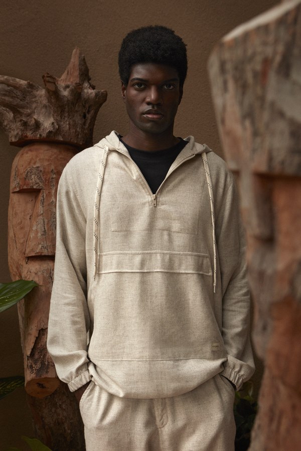 Foto de divulgação da nova coleção de roupas da marca Dafiti. Na foto, modelo posa com itens da etiqueta. É um homem jovem, negro, com cabelo curto, que veste camiseta preta e conjunto de casaco e calça bege, no tecido de linho cru.