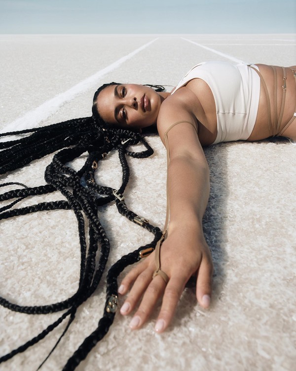 Campanha de divulgação da coleção da Nike em parceria com o estilista francês Simon Jacquemus. Na foto, a cantora Jorja Smith, uma mulher negra com cabelos longos e trançados, está deitada no chão de areia branca. Ela usa um top bege da coleção com tiras marrons amarradas pela barriga.