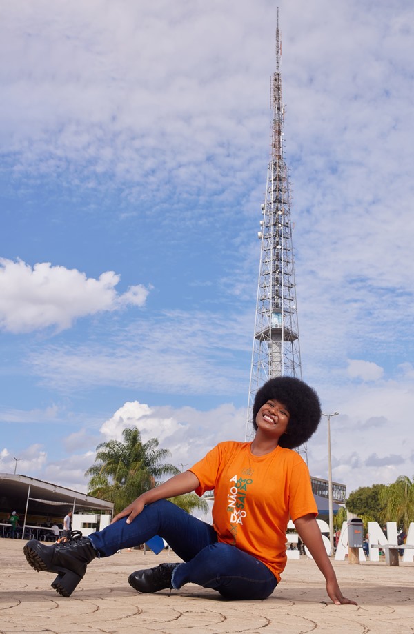 Ester Alexandre, vencedora do concurso Top Cufa DF 2022, uma menina jovem negra com cabelo cacheado posando para foto de calça jeans e camiseta laranja