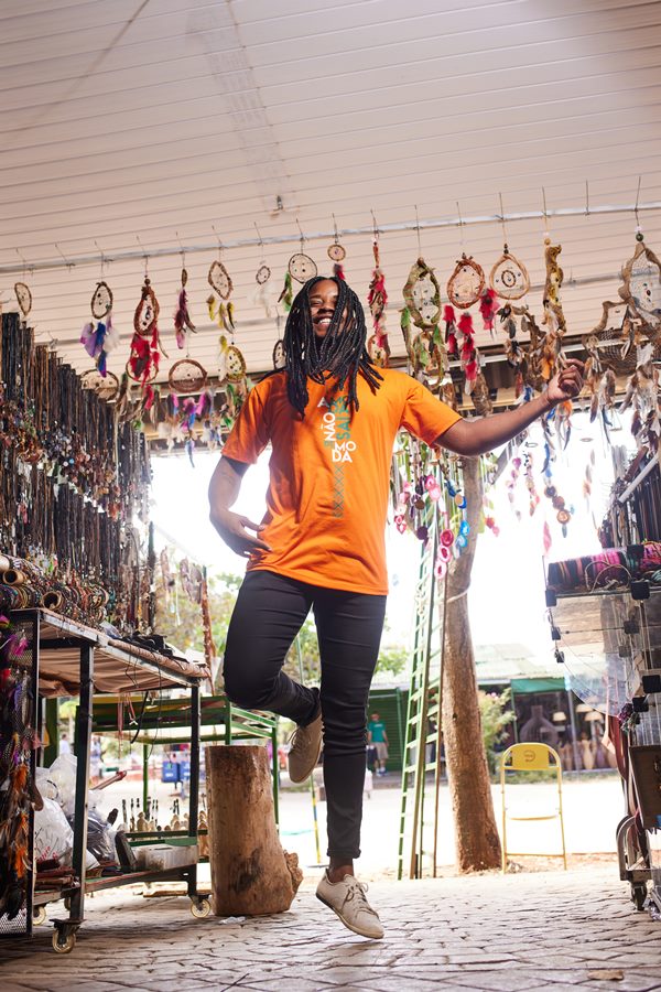 Jeferson Patrocínio Monteiro, vencedor do concurso Top Cufa DF 2022, um menino jovem negro com cabelo trançado e longo posando para foto de calça jeans e camiseta laranja