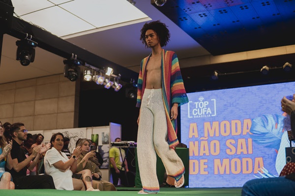 Evento Top Cufa, um concurso de beleza. Na foto, um homem negro, jovem, com cabelo cacheado desfila na passarela usando uma calça branca de crochê e um kimono colorido, também de crochê