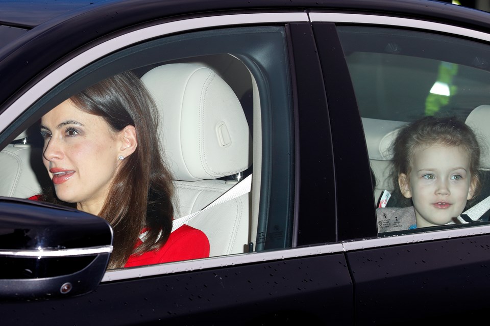 Foto colorida. Muler com roupa vermelha dentro de um carro e no banco de trás, há uma menina