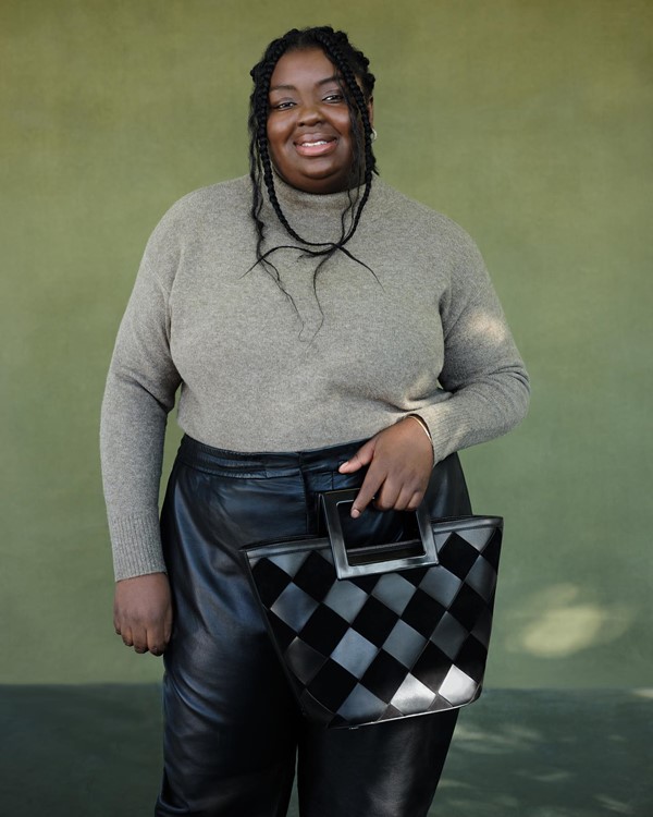 Mulher negra e jovem posando para foto em um fundo cinza. Ela tem um cabelo longo trançado e usa um casaco suéter cinza, calça de couro e uma bolsa preta