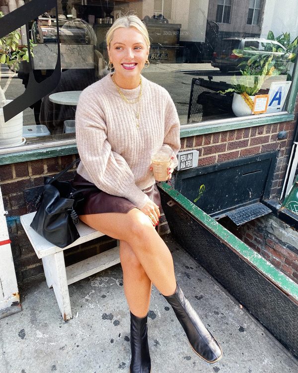 Uma mulher branca, com cabelo liso e louro, posando para foto sentada no banco externo de um restaurante. Ela usa um suéter bege de tricô, uma saia marrom, bota de couro preta e, ao seu lado no banco, é possível ver sua bolsa, também preta e de couro.