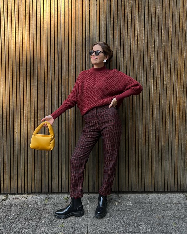 Mulher branca, com cabelo castanho amarrado, posando para foto em fundo marrom. Ela veste um suéter vinho, calça marrom e uma bolsinha amarela.