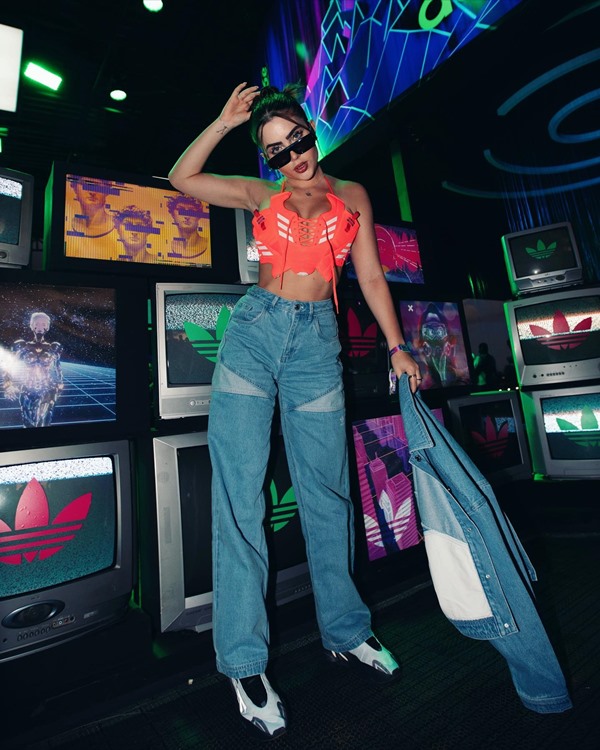 A influenciadora e ex-BBB Jade Picon, uma mulher jovem, branca e de cabelos lisos e castanhos, posando para foto em um cenário que lembra um fliperama de videogames. Ela usa uma calça jeans, um top laranja, óculos escuros e tênis preto da Nike