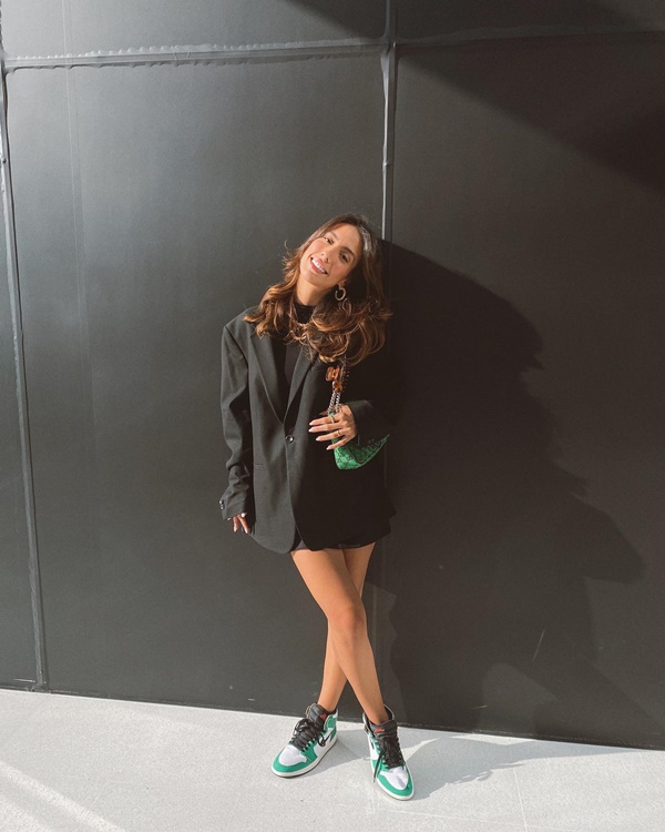 A influenciadora e ex-participante do reality show De Férias com o Ex, Gabi Brandt, posando para foto em uma parede preta. Ela é uma mulher branca com cabelos ondulados e cumpridos e veste um blazer preto, uma bolsa verde e um tênis Air Jordan, da Nike, nas cores preta, branca e verde.