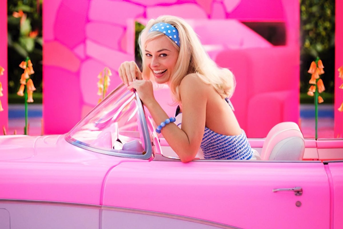 A atriz Margot Robbie, uma mulher branca, jovem e loura, gravando o filme Barbie. Ela está em um carro conversível rosa e usa um top e uma faixa na cabeça, ambos azuis com estampa de bolinha branca.