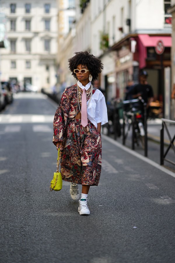 Mulher jovem e negra, com cabelo preto, crespo e curto, andando pelas ruas de Paris durante a Semana de Moda de Alta-Costura. Ela usa um óculos escuro bege, um conjunto de blusa e calça estampado vinho e azul, uma camisa branca de botão, gravata rosa clara e um tênis branco com meia branca.