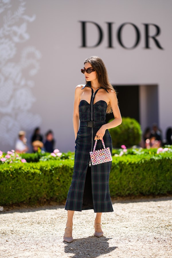 Mulher branca e jovem, de cabelo liso e castanho, posando para foto na porta do desfile da Dior durante a Semana de Alta-Costura. Ela usa um vestido longo preto com estampa xadrez, com alça amarrada no pescoço, óculos escuros, bolsa de miçangas brancas e uma sandália bege.