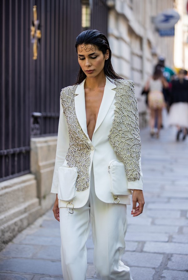 Mulher branca e jovem, de cabelo liso penteado para trás, nas ruas de Paris durante a Semana de Alta-Costura. Ela usa uma calça de alfaiataria branca e um blazer, também branco, mas com aplicações de brilhos.