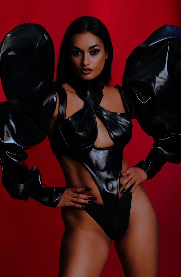 Modelo posando para a campanha do estilista Leonardo Loreto em um fundo vermelho. É uma mulher jovem e morena, de cabelo preto e liso, que veste um top preto estilo corset com estruturas que lembram asas e uma calcinha. Todas as peças são pretas.