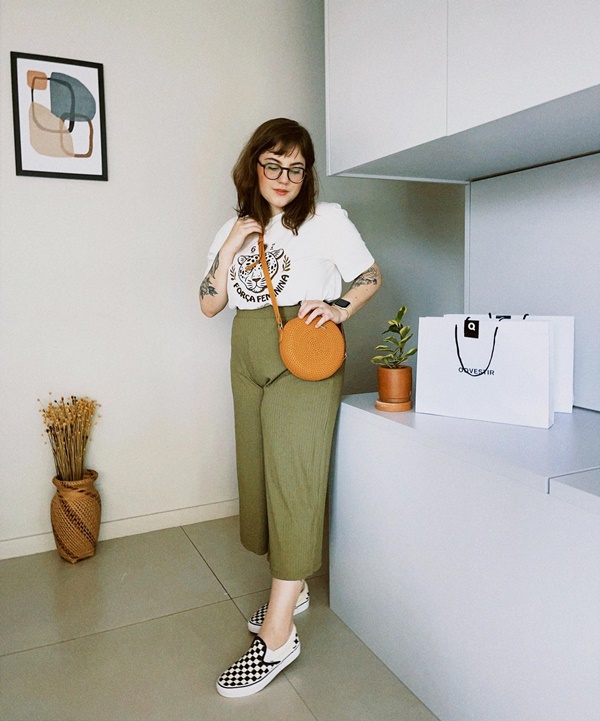 A influenciadora digital brasiliense Nanna Fernandes, uma mulher branca, jovem, com cabelos lisos e com tatuagens pelo corpo. Ela está posando para foto na sala de casa e usa uma camiseta branca, calça verde militar, tênis preto e branco e uma bolsa caramelo.