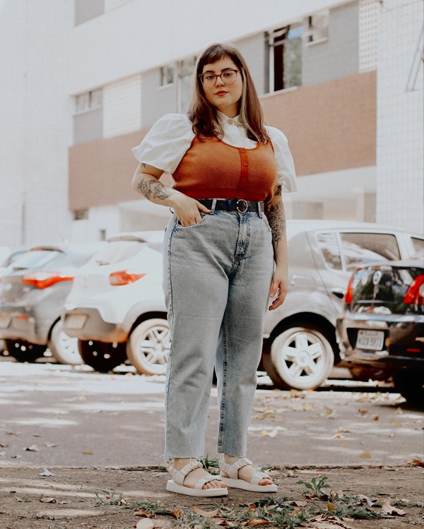 A influenciadora digital brasiliense Nanna Fernandes, uma mulher branca, jovem, com cabelos lisos e com tatuagens pelo corpo. Ela está posando para foto no estacionamento de um prédio residencia e usa uma blusa branca de manga bufantes, um colete marrom de tricô por cima, calça jeans clara e uma sandália branca.